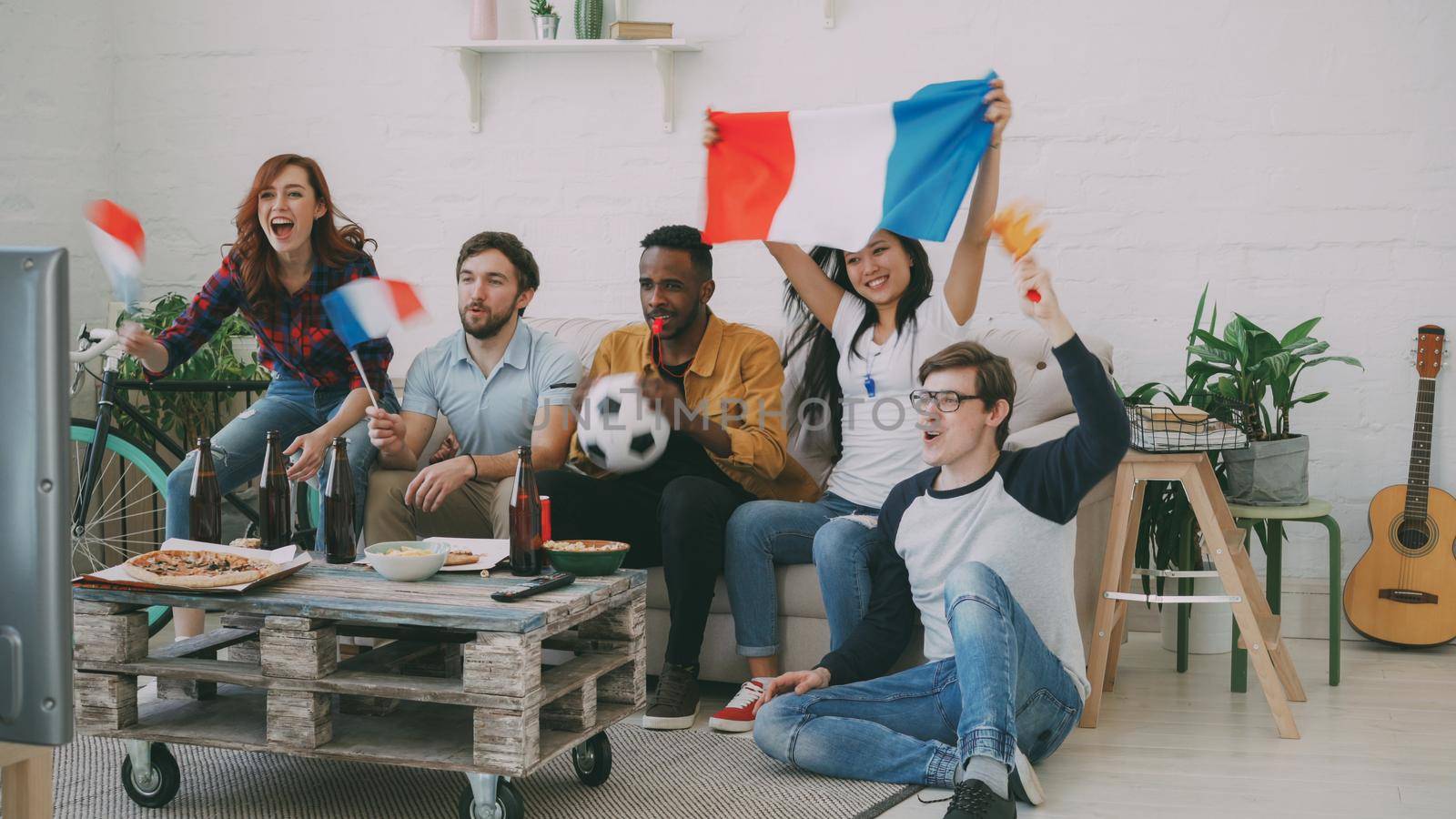 Multi-ethnic group of friends sports fans with French flags watching football championship on TV together at home indoors and cheering up favourite team