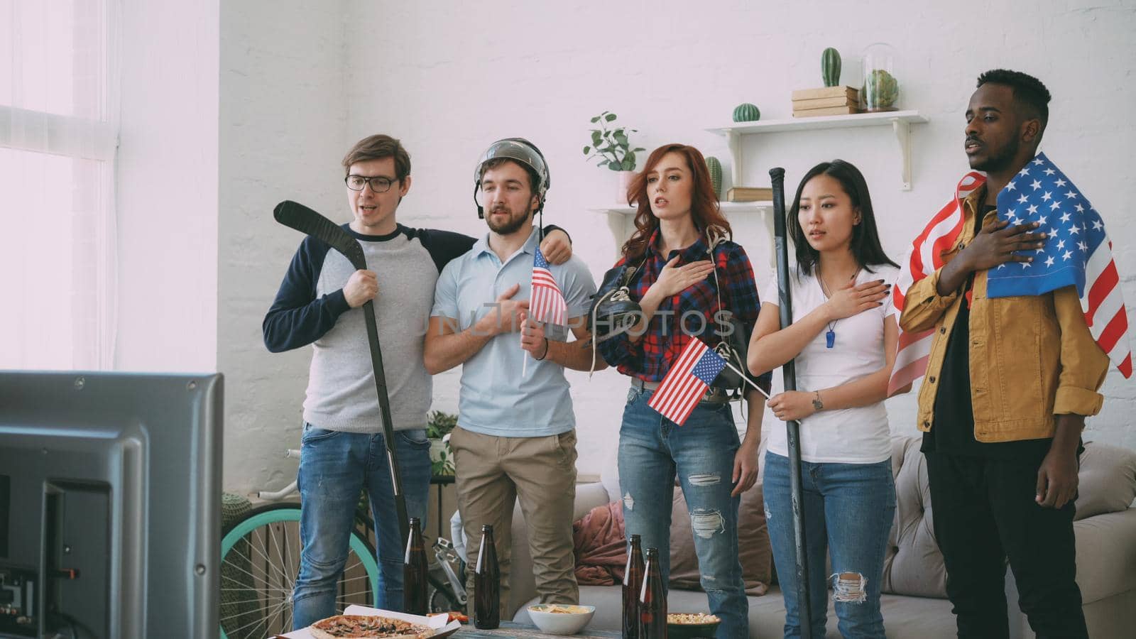 Multi ethnic group of friends sport fans listening and singing national USA anthem before watching sports championship on TV together at home indoors