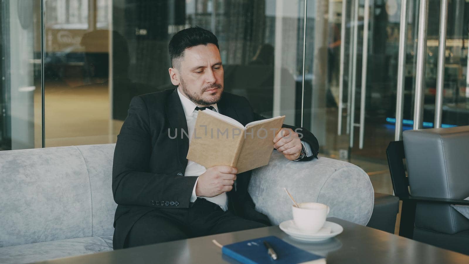 Smart Caucasian businessman in formal clothes reading text book with and drinking coffee in classy cafe at lunch break by silverkblack