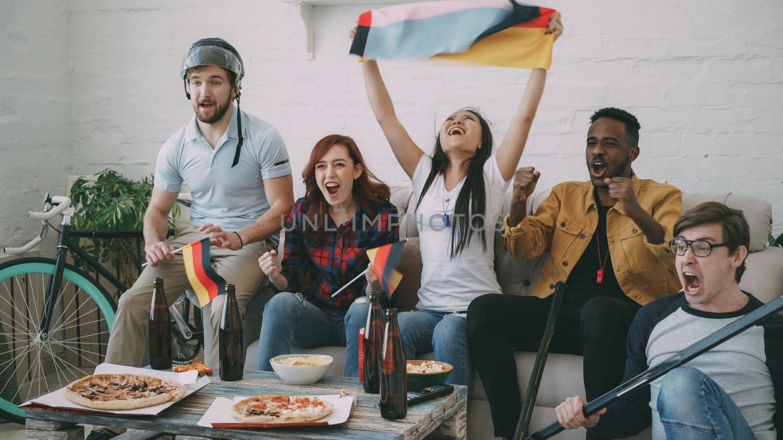 Multi ethnic group of friends sports fans with German national flags watching hockey championship on TV together cheering up their favourite team at home indoors
