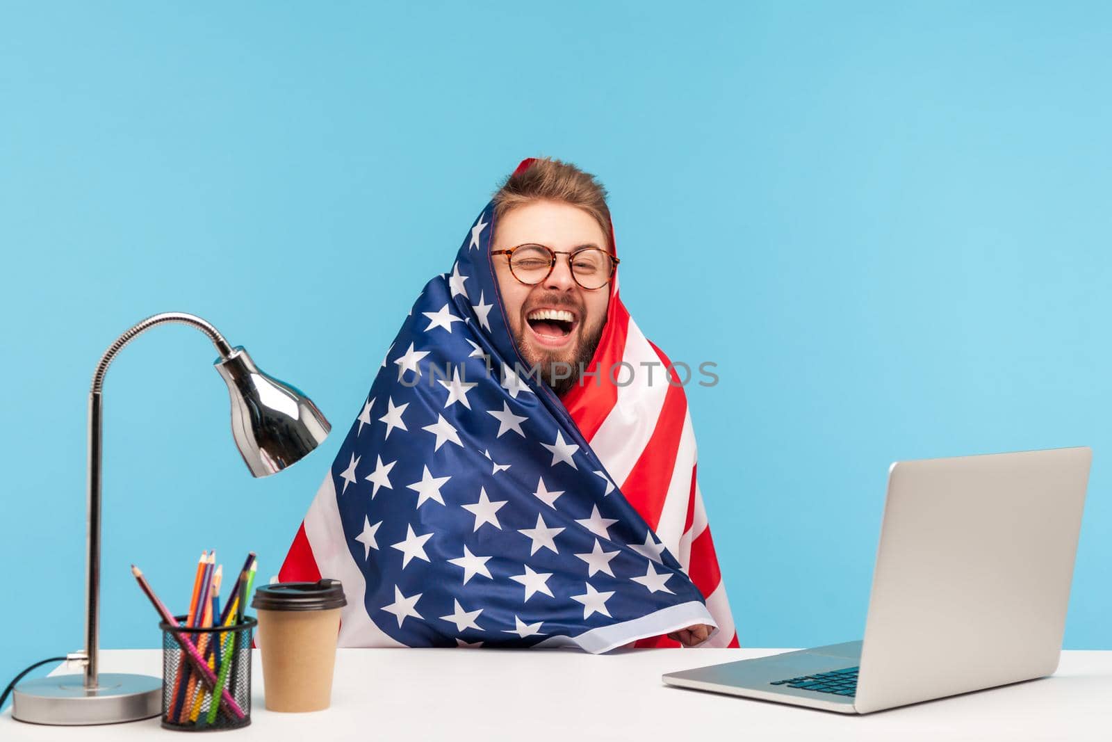 Extremely happy funny man employee sitting wrapped in American flag and shouting for joy in office workplace, celebrating labor day or US Independence day 4th of july, government employment support.