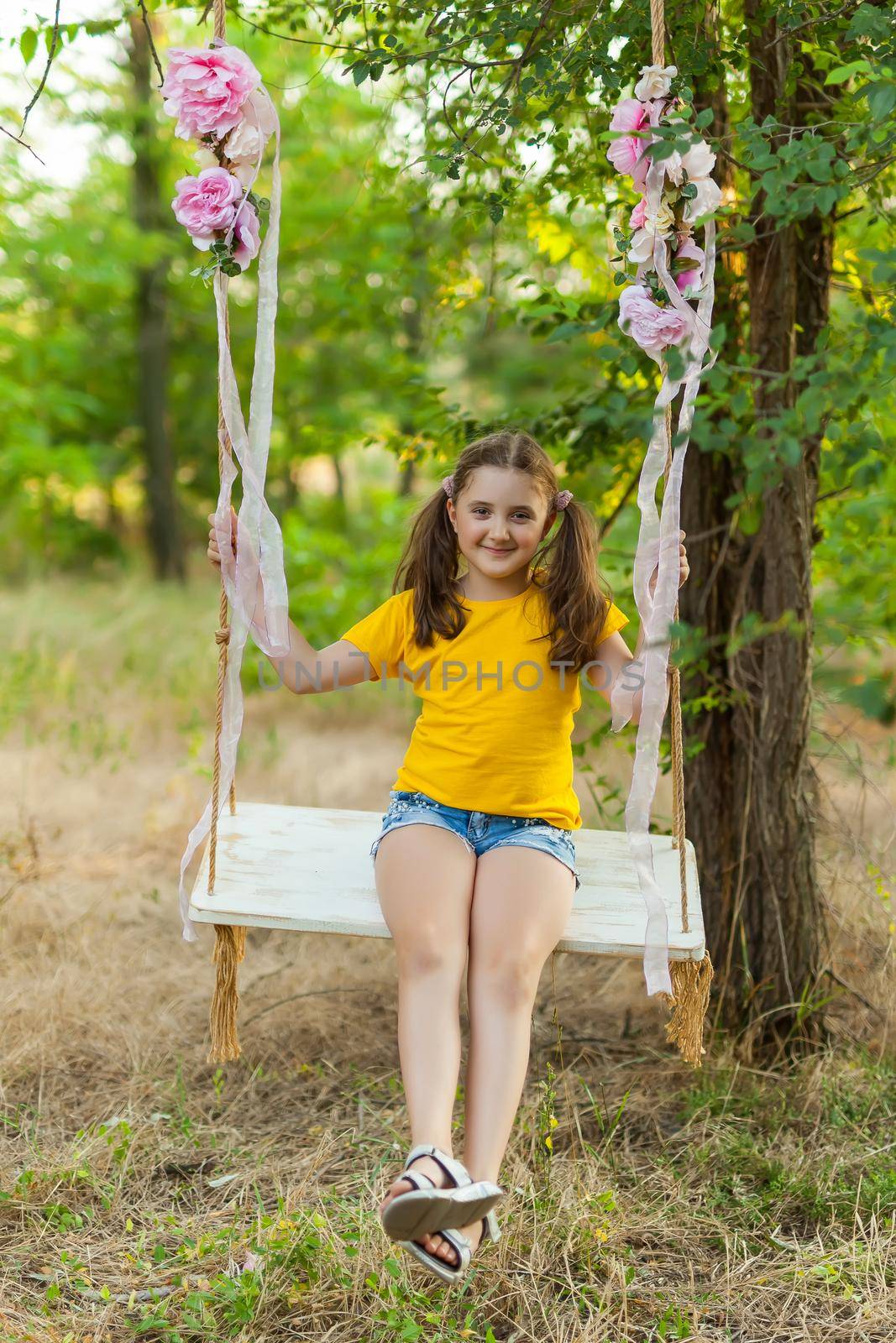Cute smiling girl in a yellow t-shirt having fun on a swing in tree forest. Sunny day. Summer outdoor activities for kids