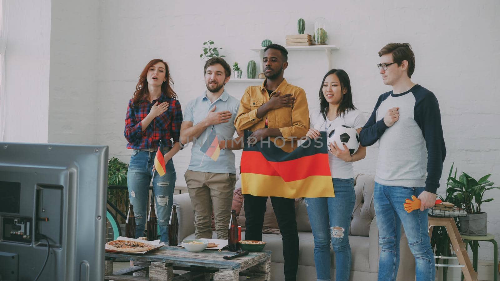 Multi-ethnic group of friends listening and singing German national anthem before watching sports championship on TV together at home by silverkblack