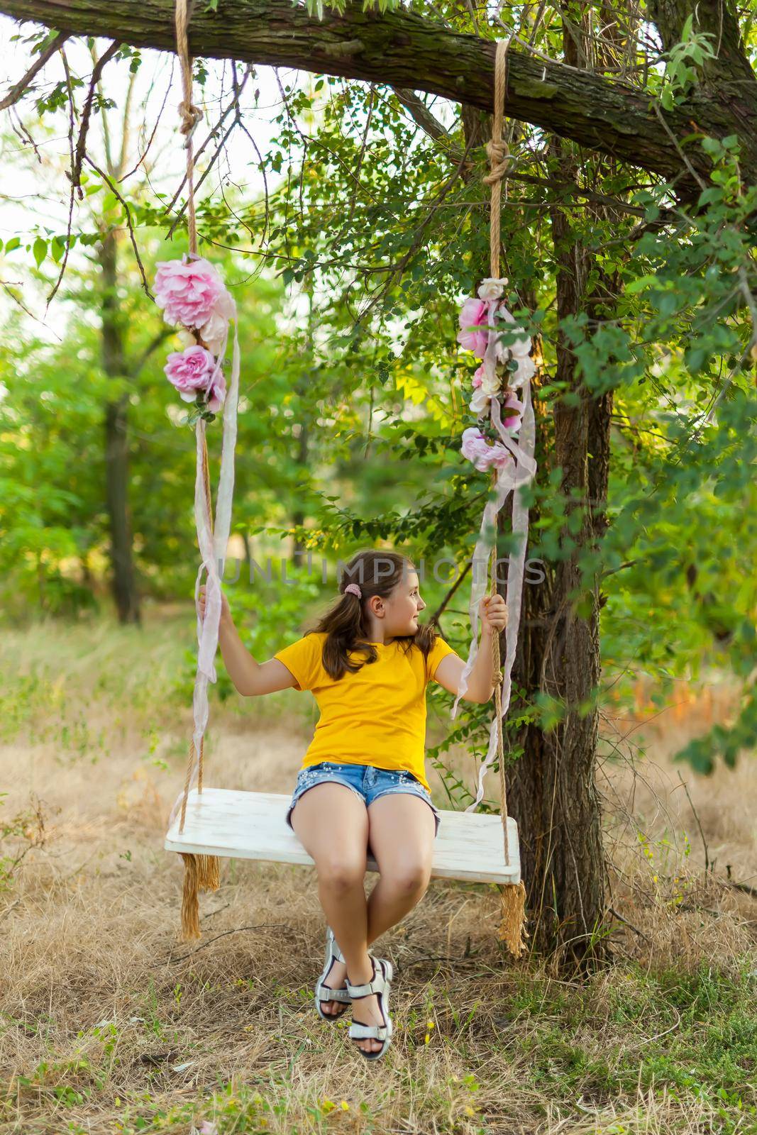 Cute smiling girl in a yellow t-shirt having fun on a swing in tree forest. Sunny day. Summer outdoor activities for kids