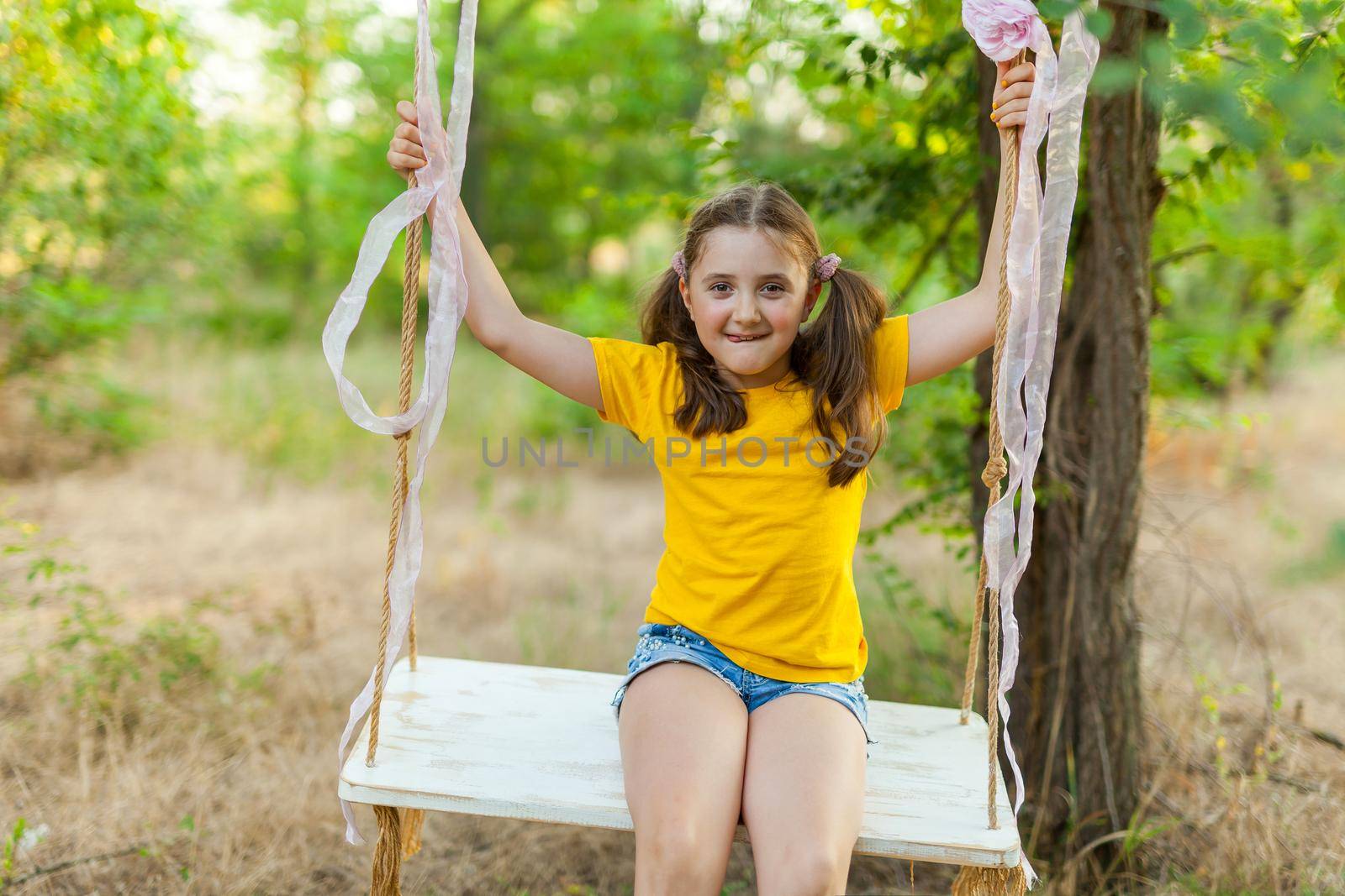 Cute smiling girl in a yellow t-shirt having fun on a swing in tree forest. Sunny day. Summer outdoor activities for kids