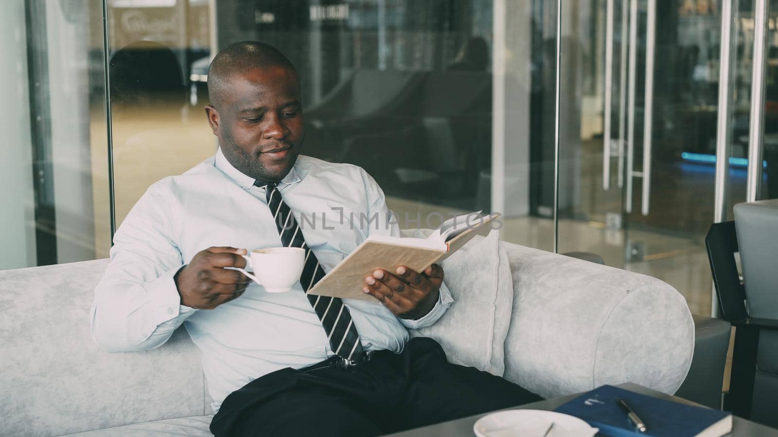 Smart African American businessman in formal clothes reading book while drinking coffee in glassy cafe at lunch break by silverkblack