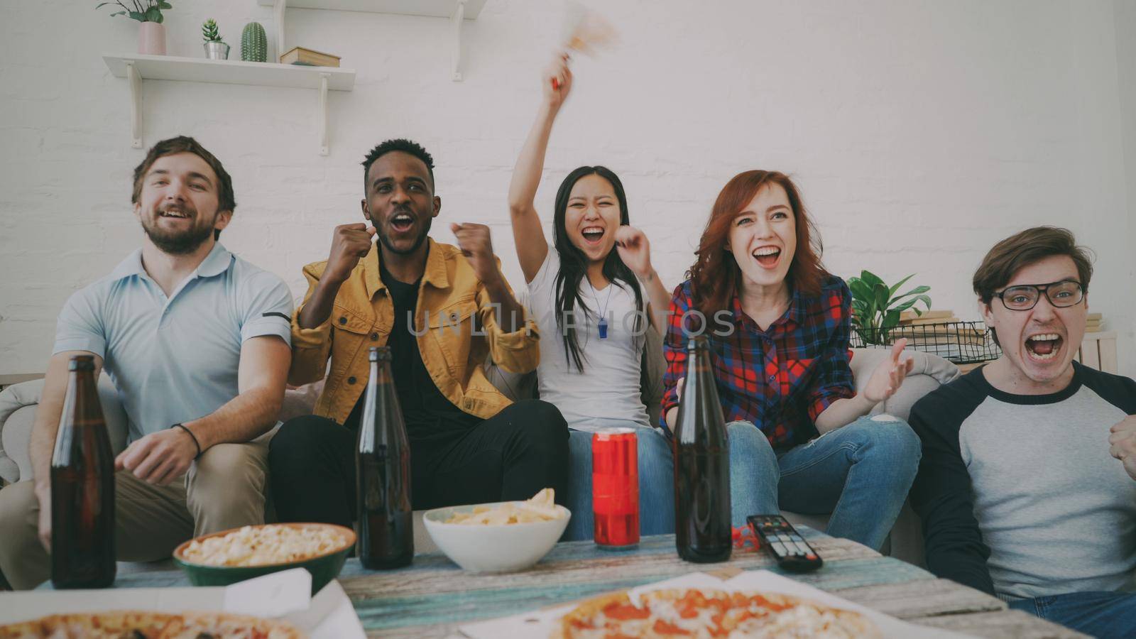 Group of young friends watching olympic games match on TV together eating snacks and drinking beer. Some of them happy with their team winning but african man disappointed by silverkblack