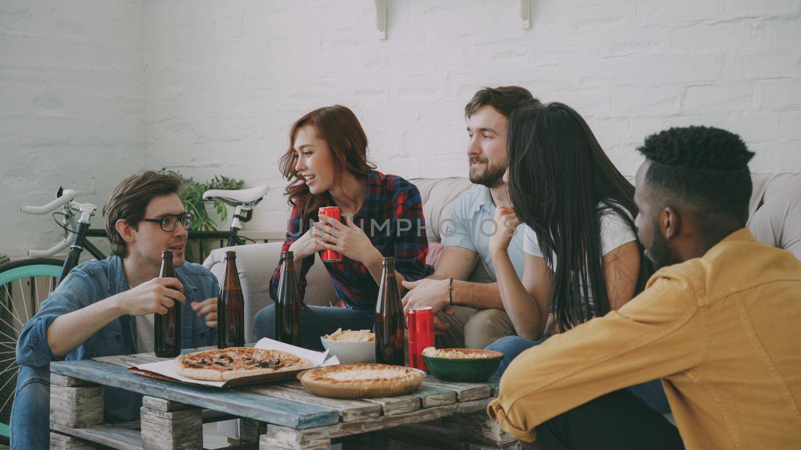 Multiracial young cheerful friends enjoying sitting together and talking each other while drinking beer and eating snacks at home by silverkblack