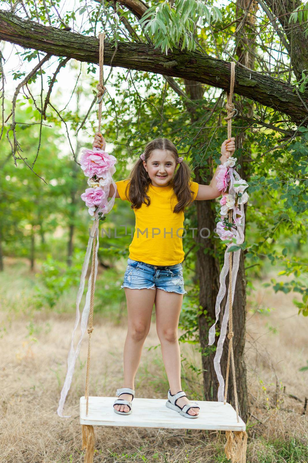 Cute smiling girl in a yellow t-shirt having fun on a swing in tree forest. Sunny day. Summer outdoor activities for kids