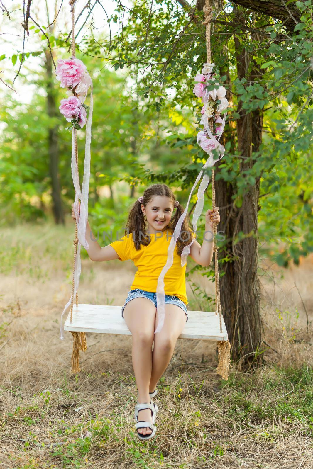 Cute smiling girl in a yellow t-shirt having fun on a swing in tree forest. Sunny day. Summer outdoor activities for kids