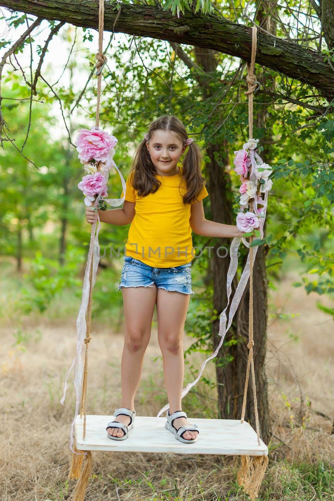 Cute smiling girl in a yellow t-shirt having fun on a swing in tree forest. Sunny day. Summer outdoor activities for kids