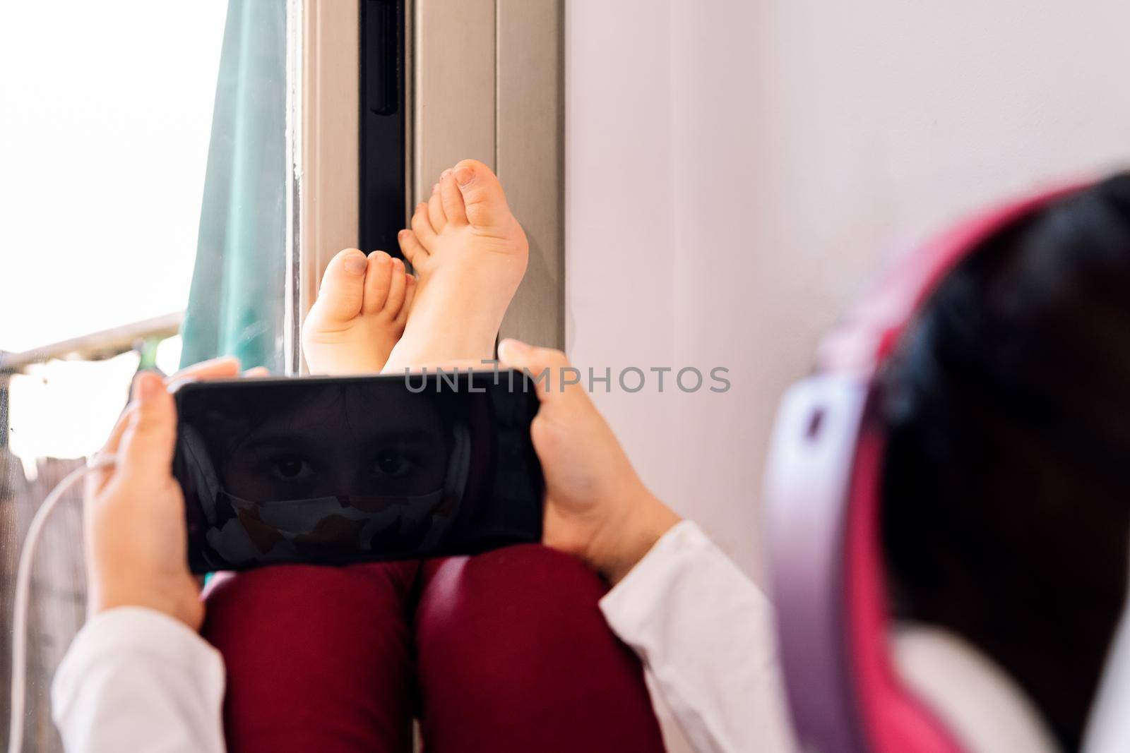 child sitting at home with a headset and protective mask reflected on her cell phone screen, coronavirus concept and communication in quarantine