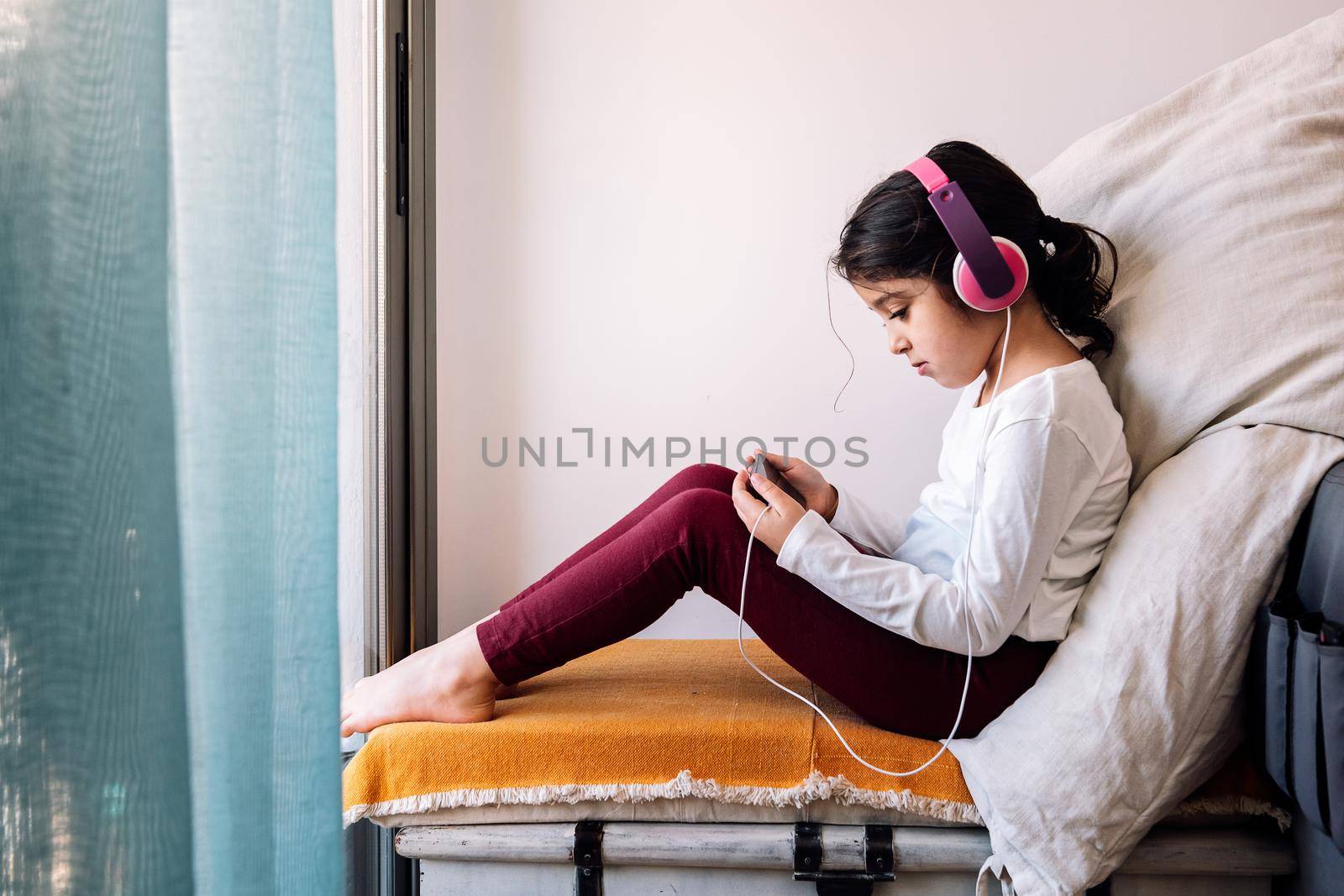little girl sitting in front of her home window with headphones watching videos on the phone, home entertainment for children concept