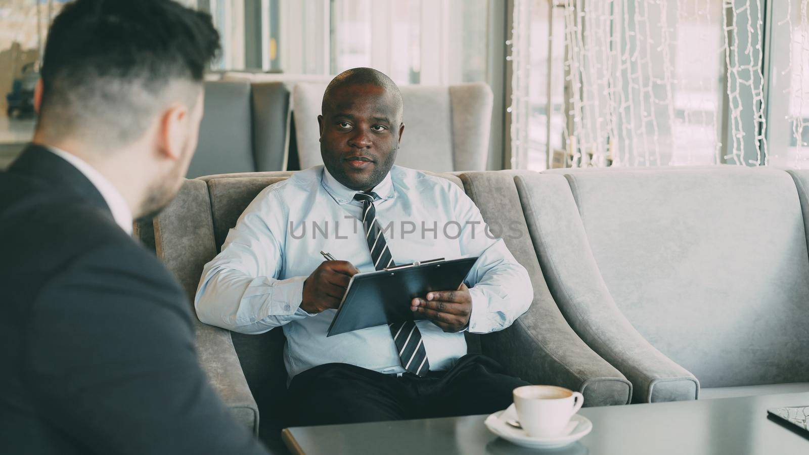 African american HR manager having job interview with young man in suit and watching his resume application in modern cafe indoors