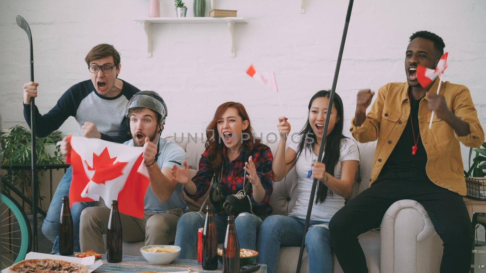 Group of young friends sports fans with Canadian national flags watching sport championship on TV together cheering up favourite team at home indoors
