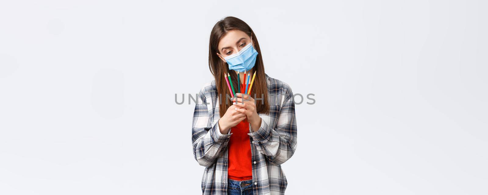 Social distancing, leisure and hobbies on covid-19 outbreak, coronavirus concept. Thoughtful cute, dreamy woman in medical mask decide which colored pencil choose, pondering what draw.
