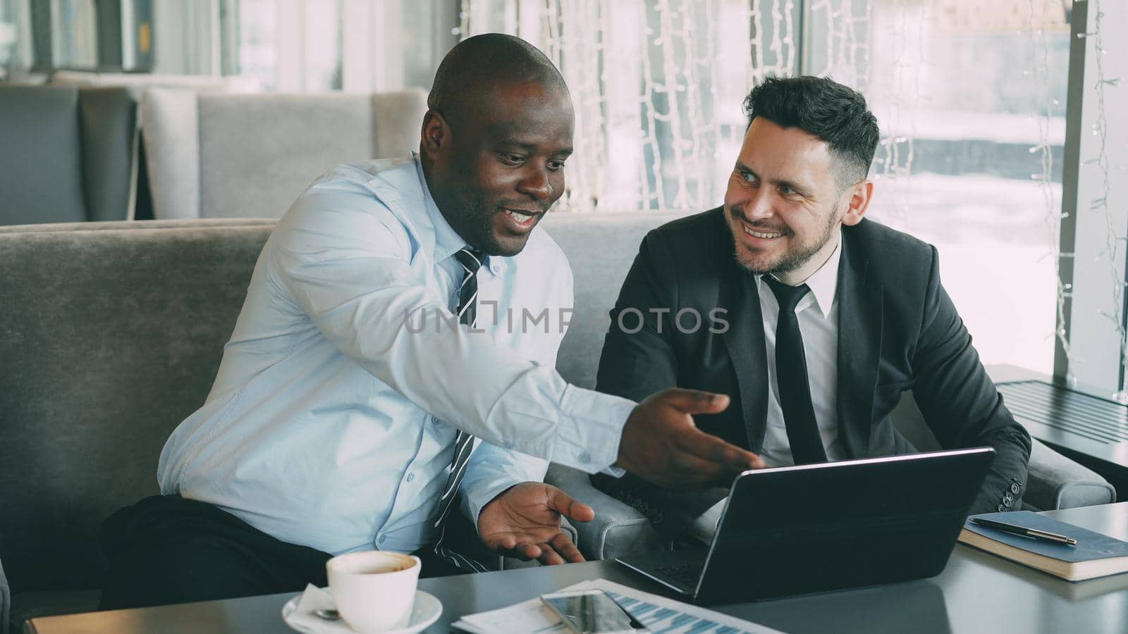 Cheerful African American businessman in formal clothes discussing business project with his caucasian colleague on his laptop in cafe by silverkblack