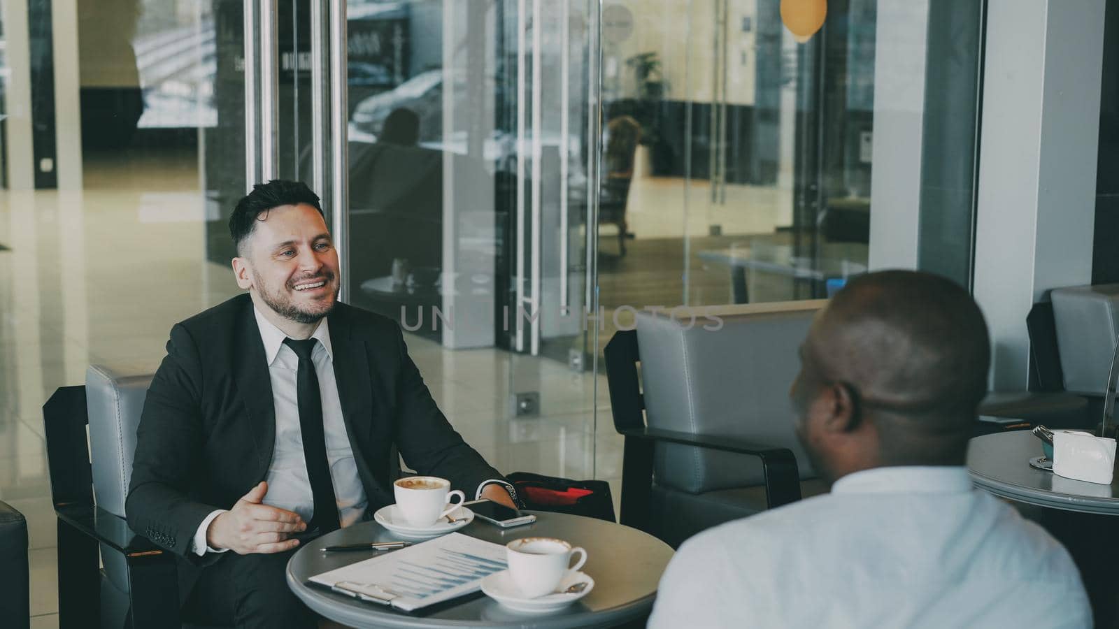Cheerful bearded Caucasian businessman in formal clothes smiling, gesticulating, drinking coffee and discussing financial report of startup with his African American partner in modern cafe