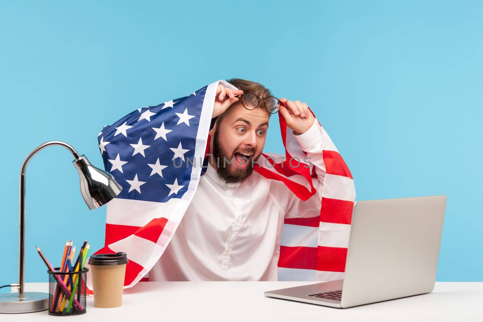 4th of july. Amazed, extremely happy businessman sitting wrapped in American flag, watching live patriotic program on laptop at workplace and shouting for joy with surprised shocked expression. indoor