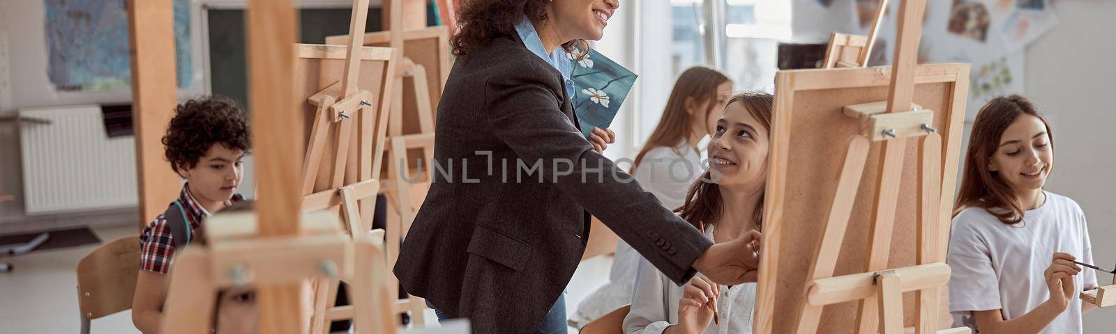 Young beautiful confident teacher is helping a kid to draw on a group lesson in a white modern minimalistic classroom by Yaroslav_astakhov