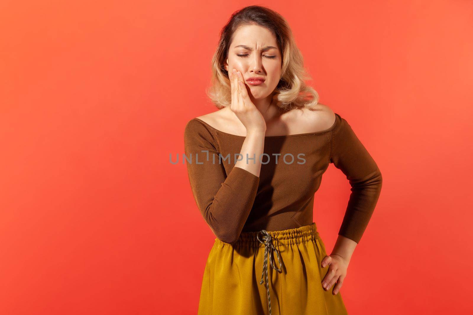 Dental problem. Unhealthy sick blonde woman touching cheek, closing eyes with expression of terrible suffer from toothache, sensitive teeth, cavities. Indoor studio shot isolated on red background