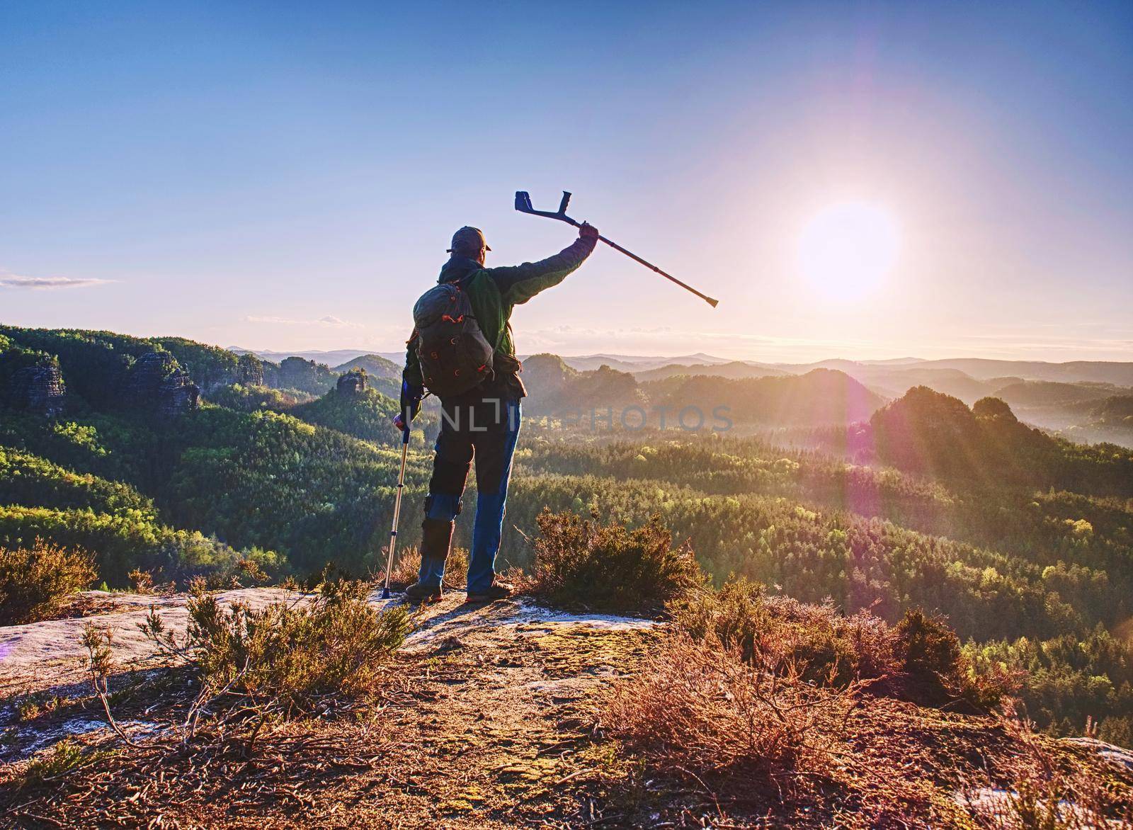 Hiker with medicine crutch achieved mountain peak. Broken leg fixed in immobilizer.  Open mountain valley bellow cliff. 