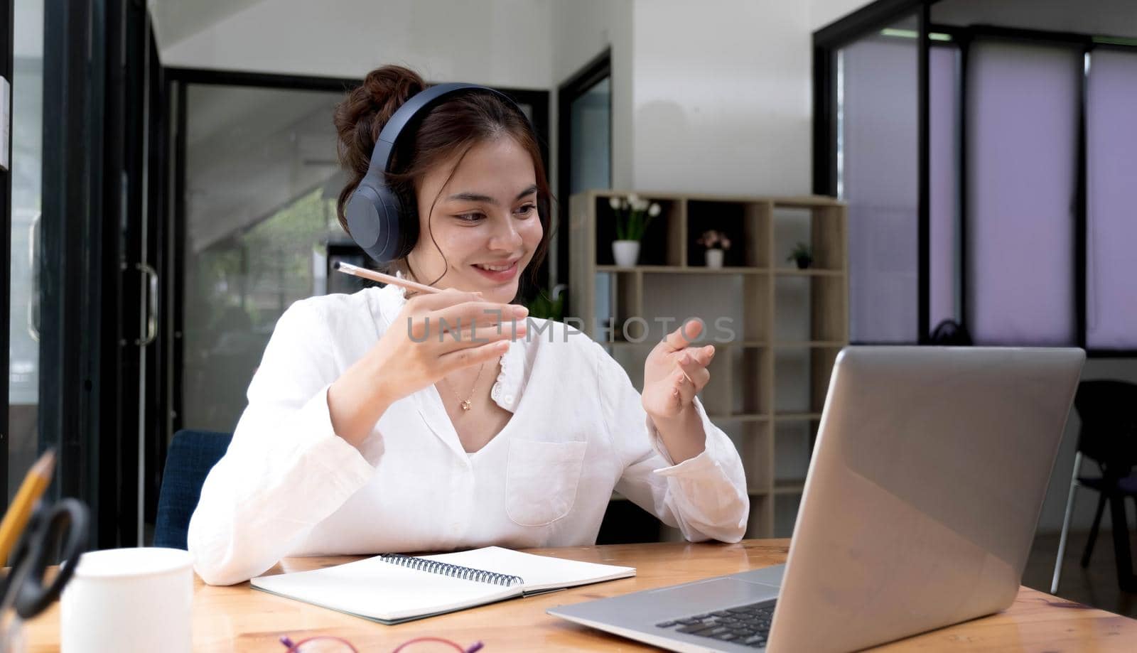 Smiling young asian woman in headset wave greet talking on webcam virtual conversation on laptop, happy female in wireless headphones speak on video call on computer, consult client online by wichayada