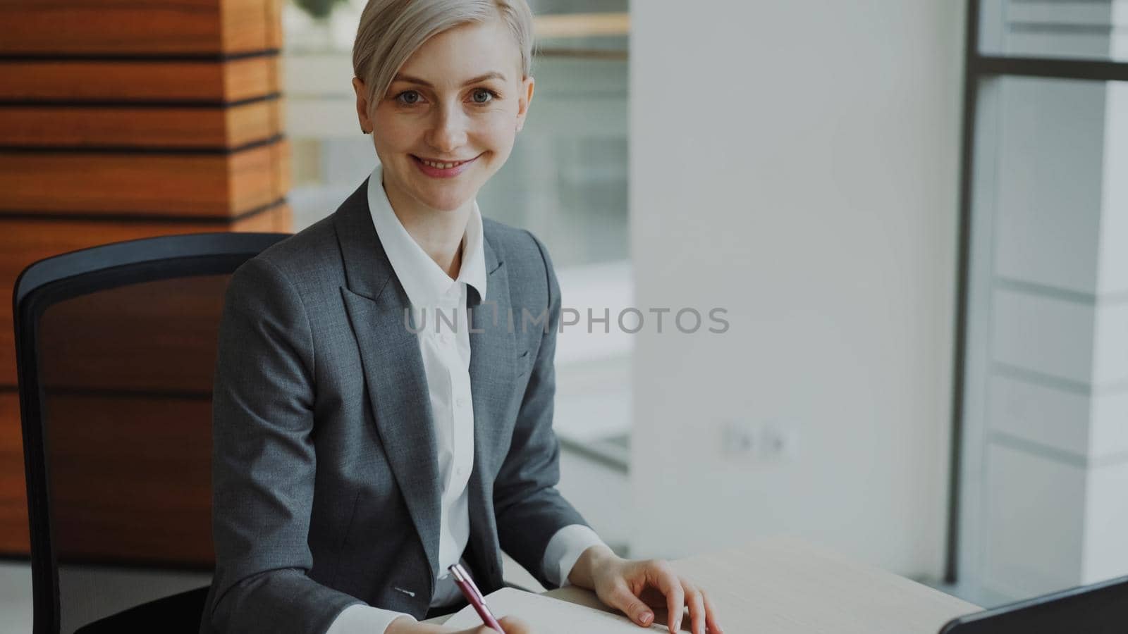 Portrait of Attractive blonde businesswoman sitting at table writing in notebook smiling into camera in modern office by silverkblack