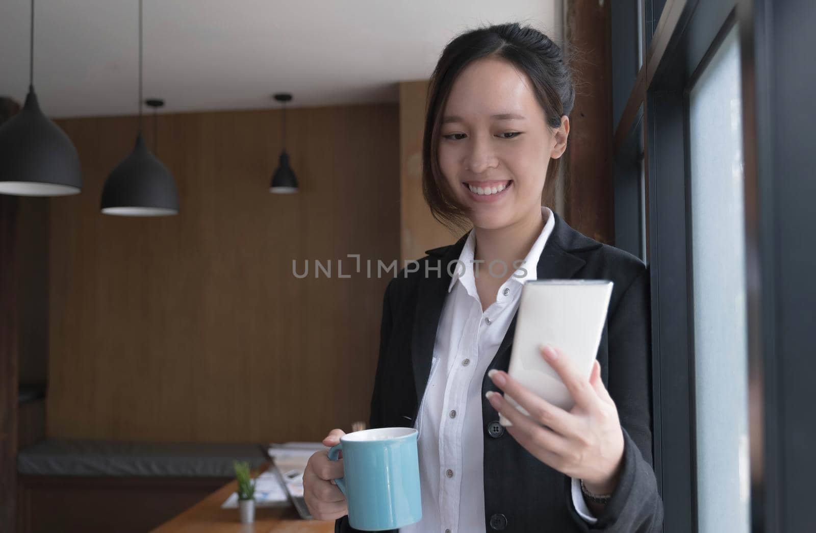 Happy young Asian businesswoman standing using smartphone and hold coffee cup at office..