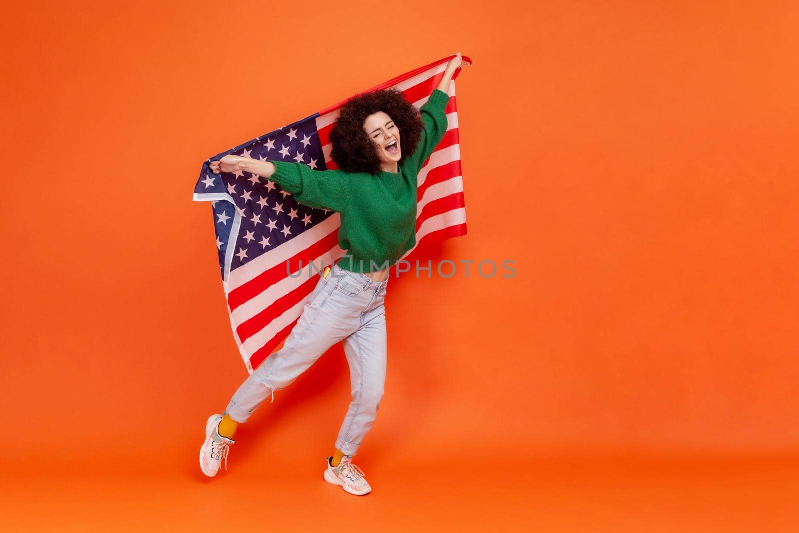 Full length photo of woman with Afro hairstyle wearing green casual style sweater raised arms, holding american flag, celebrating national holiday. Indoor studio shot isolated on orange background.