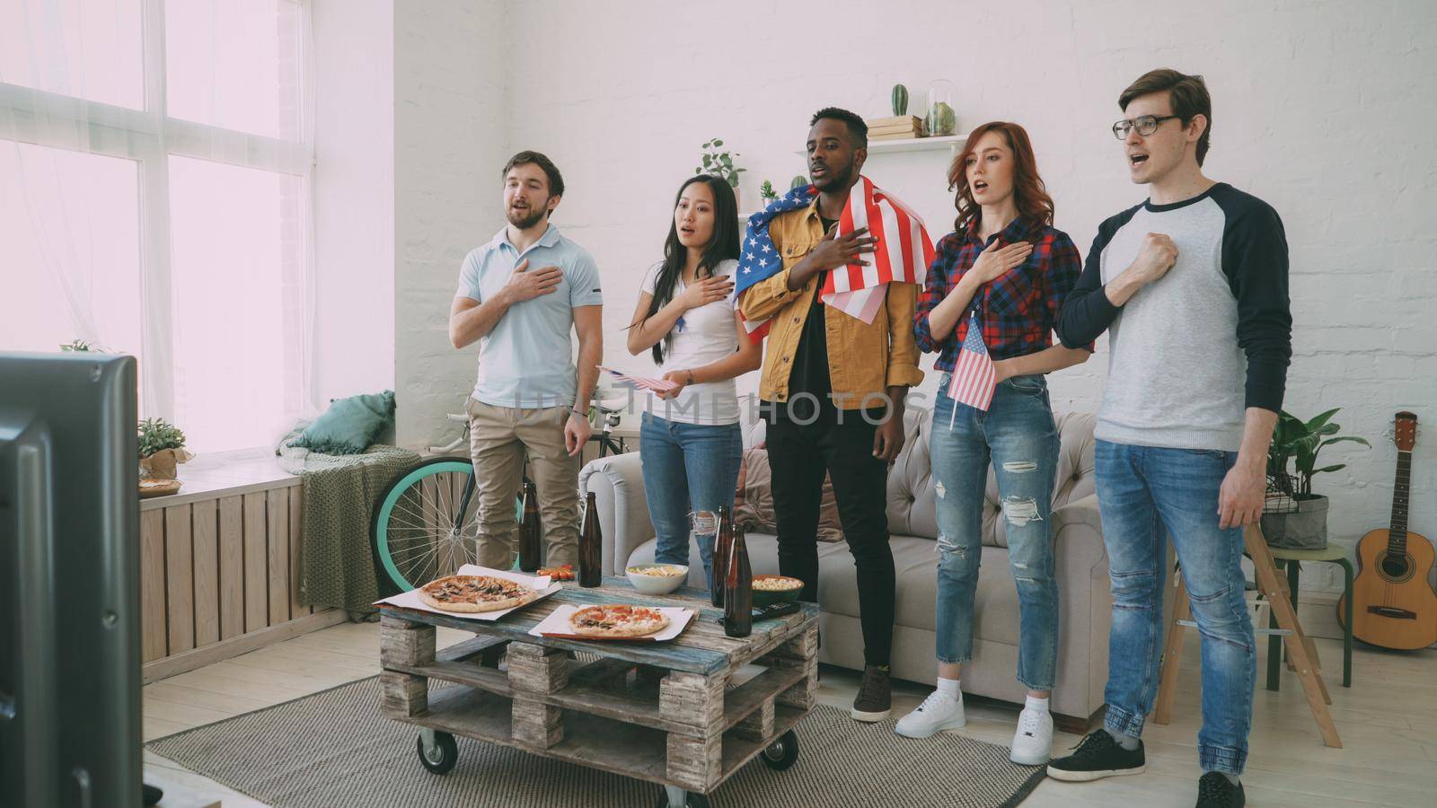 Multi ethnic group of friends sport fans singing national USA anthem before watching sports championship on TV together at home by silverkblack