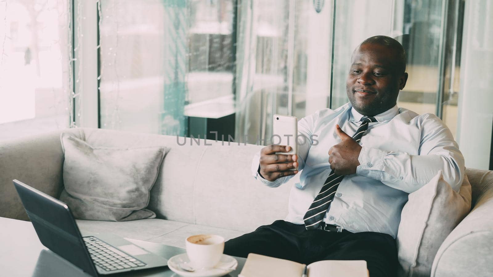 Happy African American businessman taking a selfie on his smartphone and showing a thumb up gesture while drinking coffee in modern cafe. by silverkblack