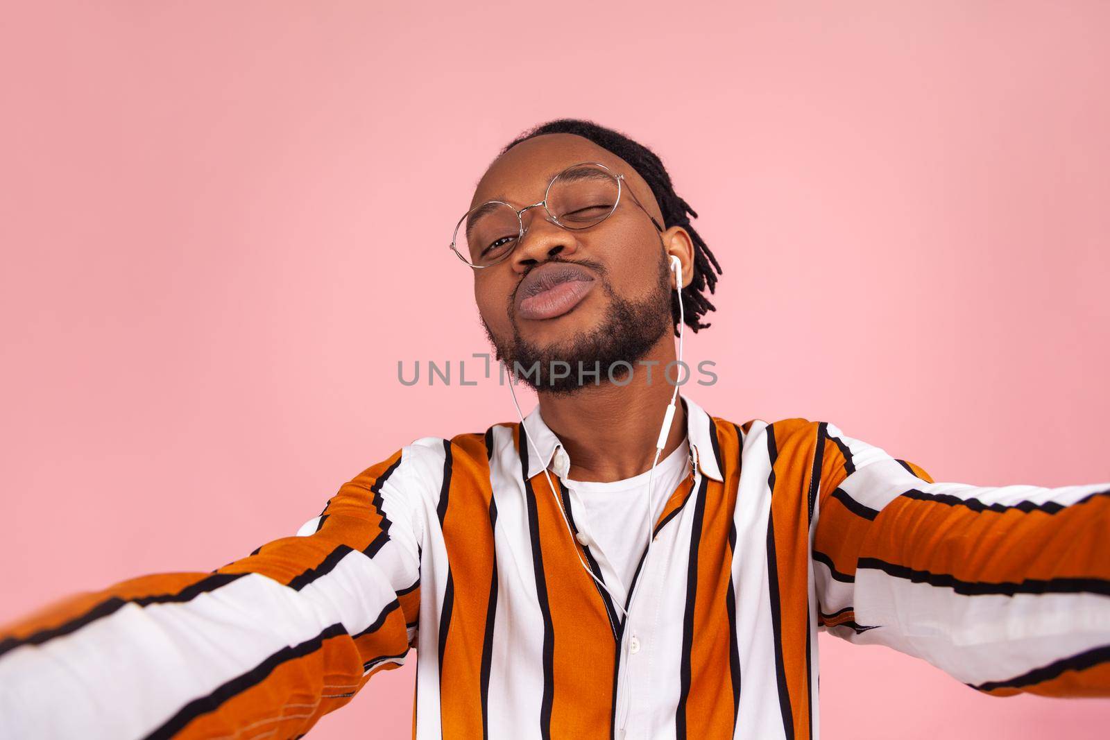 Funny bearded afro-american man blogger with dreadlocks in eyeglasses and striped shirt sending air kiss looking at selfie camera, video call. Indoor studio shot isolated on pink background