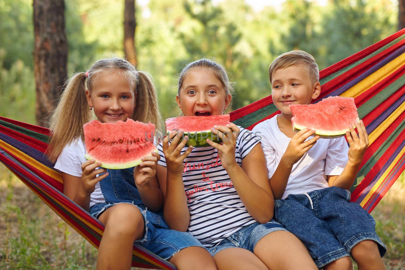 three cheerful children eat watermelon and joke, outdoor, sitting on a colorful hammock. Summer fun and leisure