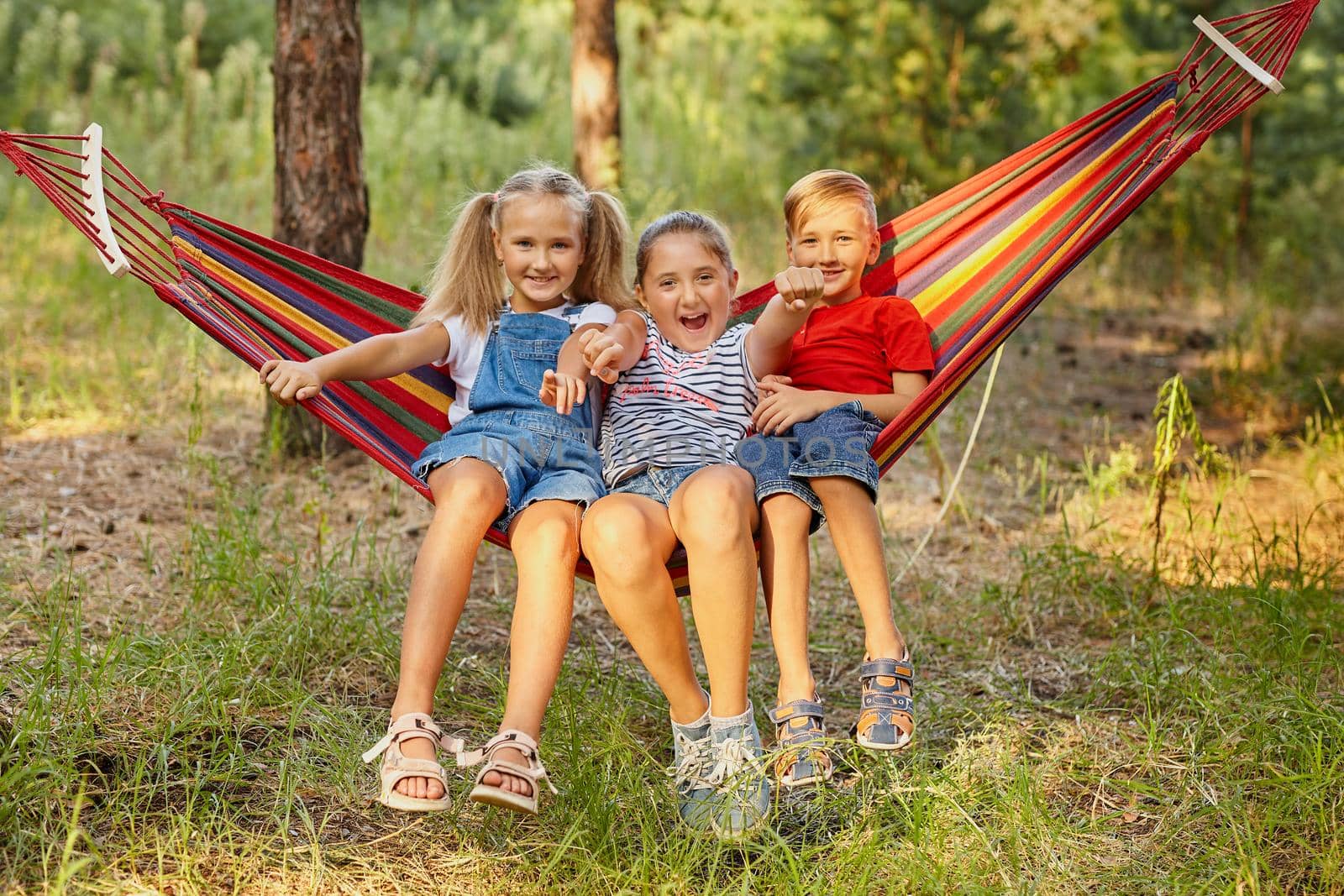 three cheerful children joke, outdoor, sitting on a colorful hammock. Summer fun and leisure