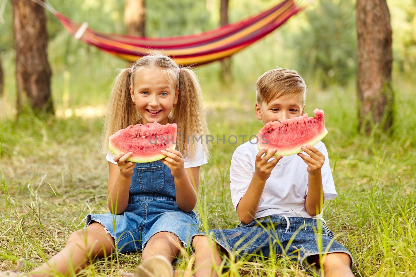 Happy Kids eating watermelon in the park. Kids eat fruit outdoors. Healthy snack for children. Little girl and boy playing in the forest biting a slice of water melon