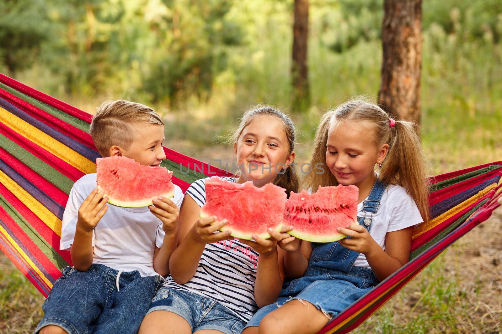 three cheerful children eat watermelon and joke, outdoor, sitting on a colorful hammock. Summer fun and leisure