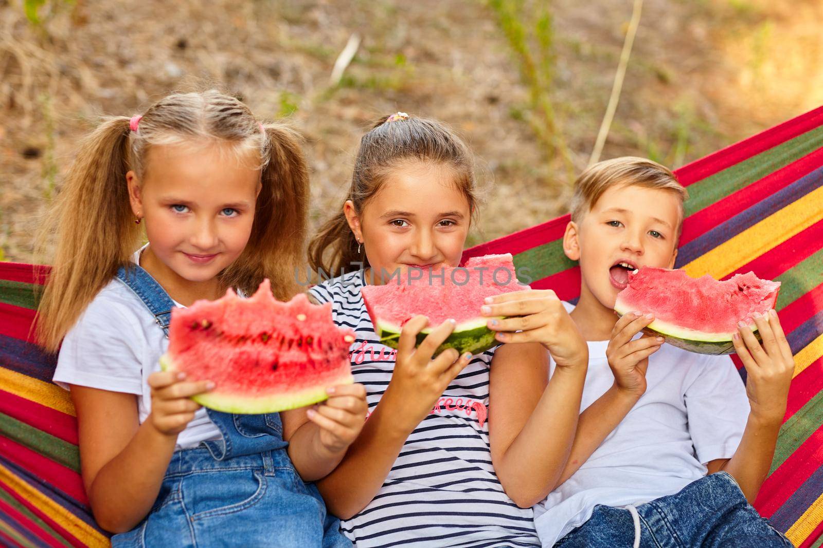 children eat watermelon and joke, outdoor, sitting on a colorful hammock. by InnaVlasova