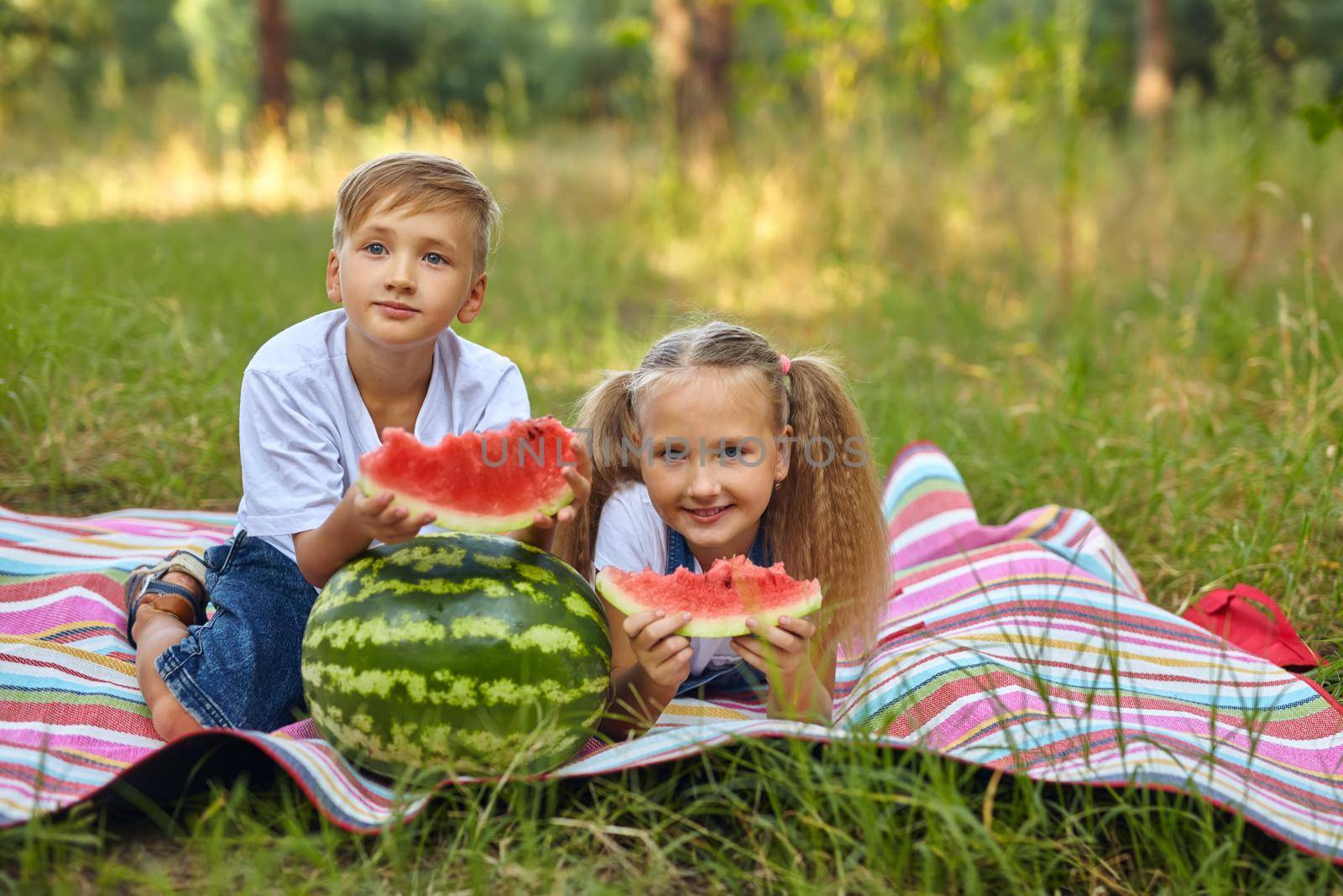 Kids eating watermelon in the park. Kids eat fruit outdoors. Healthy snack for children. Little girl and boy playing in the forest biting a slice of water melon