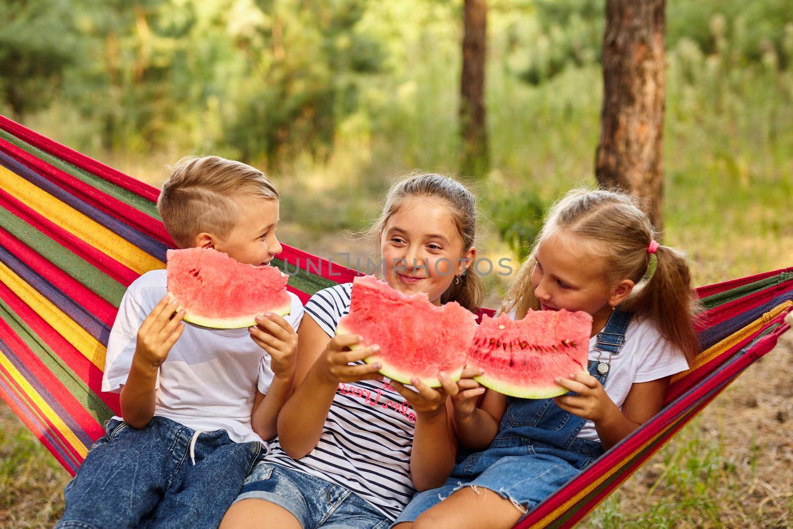 children eat watermelon and joke, outdoor, sitting on a colorful hammock. by InnaVlasova