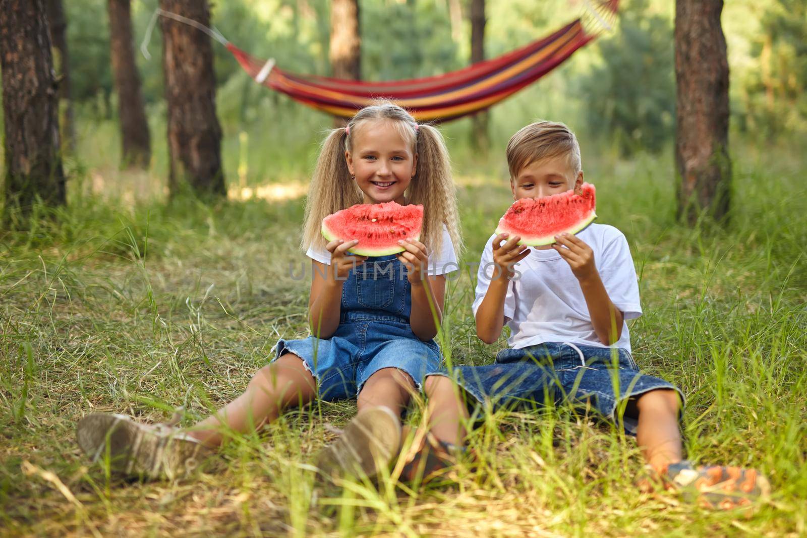 Kids eating watermelon in the park. Kids eat fruit outdoors. Healthy snack for children. Little girl and boy playing in the forest biting a slice of water melon
