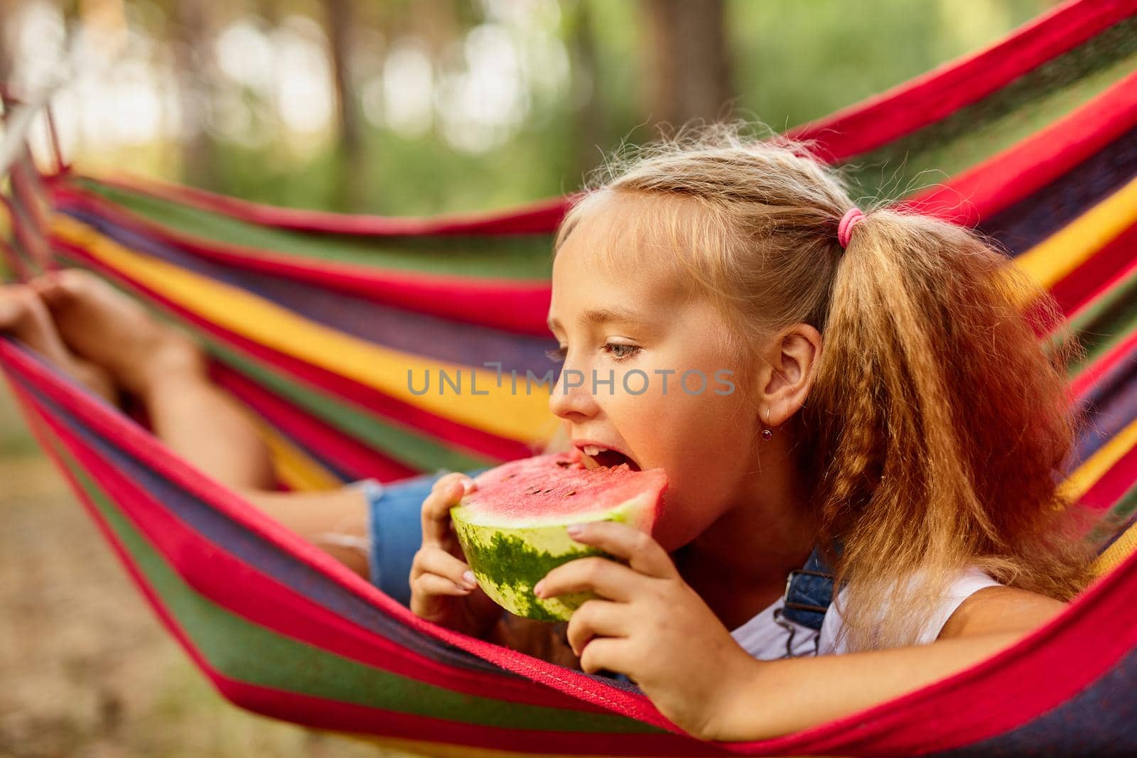 Cute little girl resting in a colored hammock in the forest and eating fresh watermelon