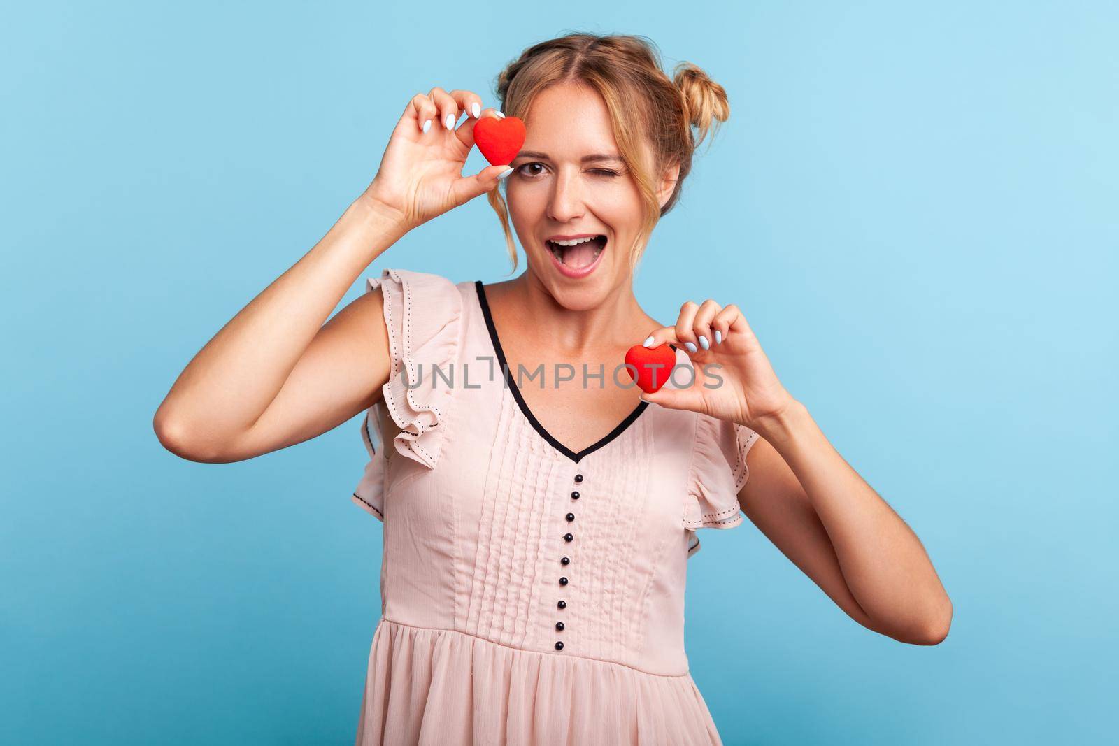 Portrait of friendly positive blonde woman in dress with two hair buns holding heart symbols in hands and winking at camera, sending love. Indoor studio shot isolated on blue background.