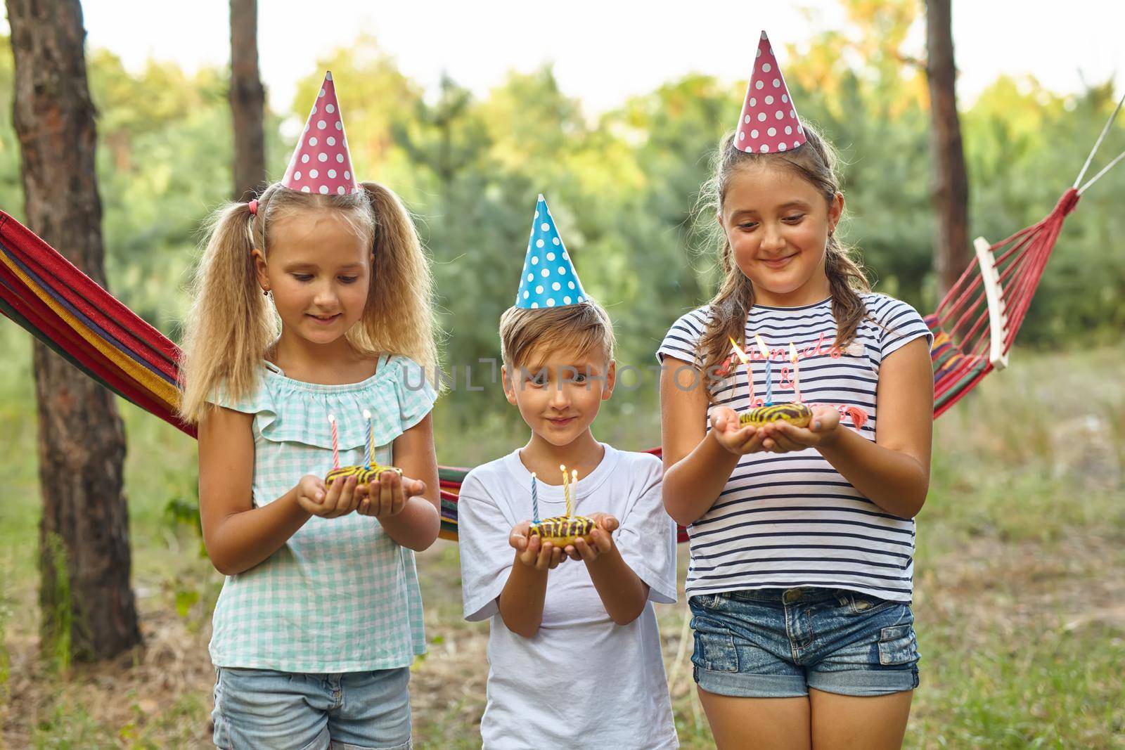 Children holding birthday cakes with burning candles. Kids party decoration and food. Boy and girls celebrating birthday in the garden with hammock. Kids with sweets.