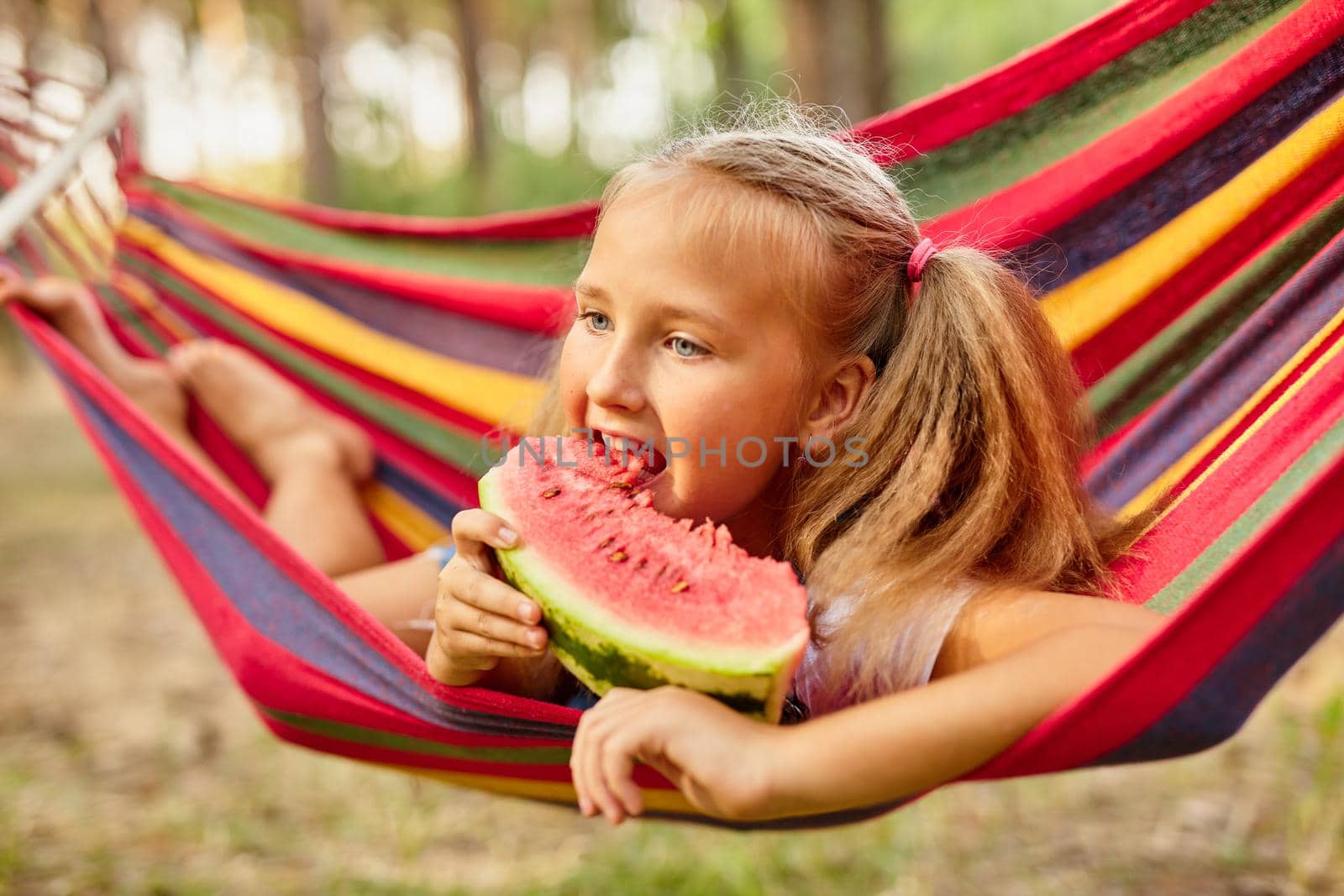 Cute little girl resting in a colored hammock in the forest and eating fresh watermelon