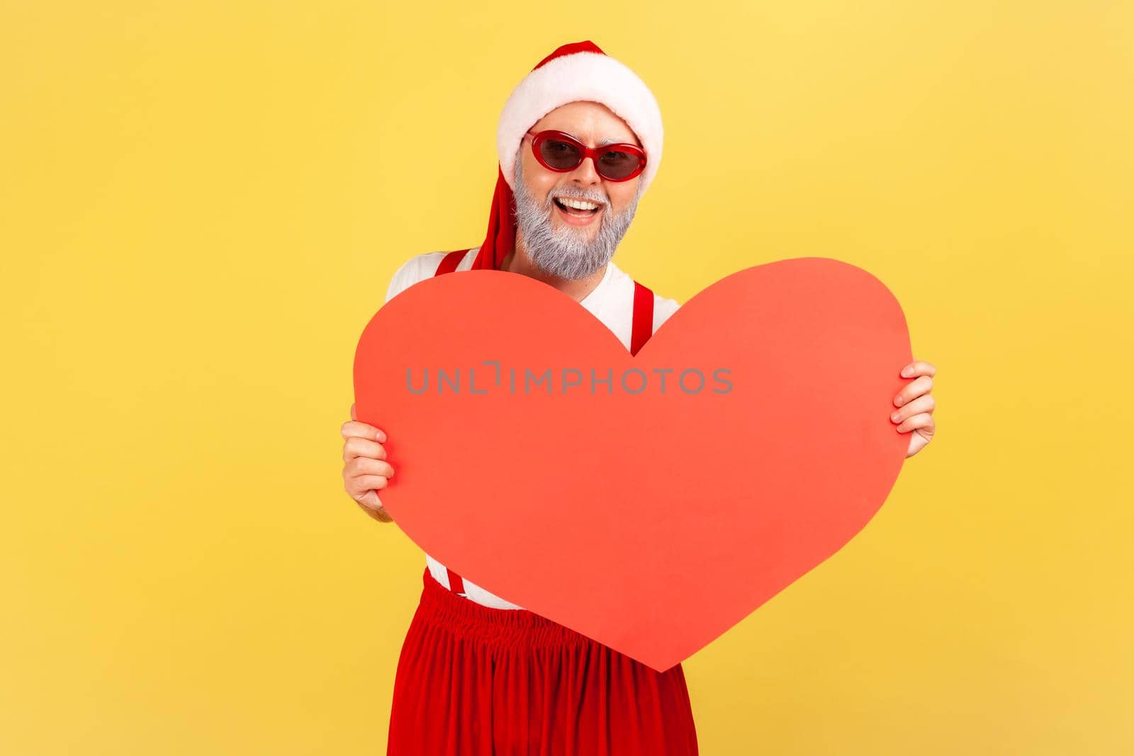 Positive cheerful elderly man in trendy sunglasses and santa claus hat holding and showing paper heart, symbol of love, winter holidays. Indoor studio shot isolated on yellow background