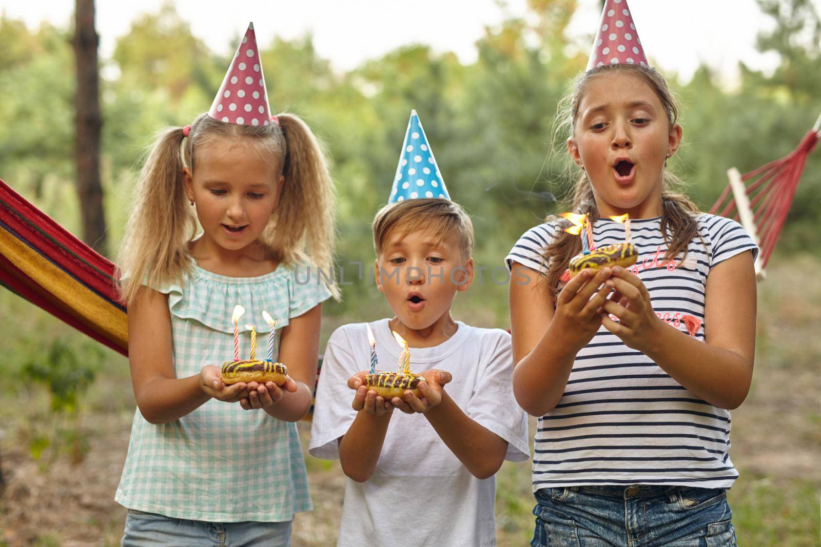 Children blow candles on birthday cake. Kids party decoration and food. Boy and girls celebrating birthday in the garden with hammock. Kids with sweets.