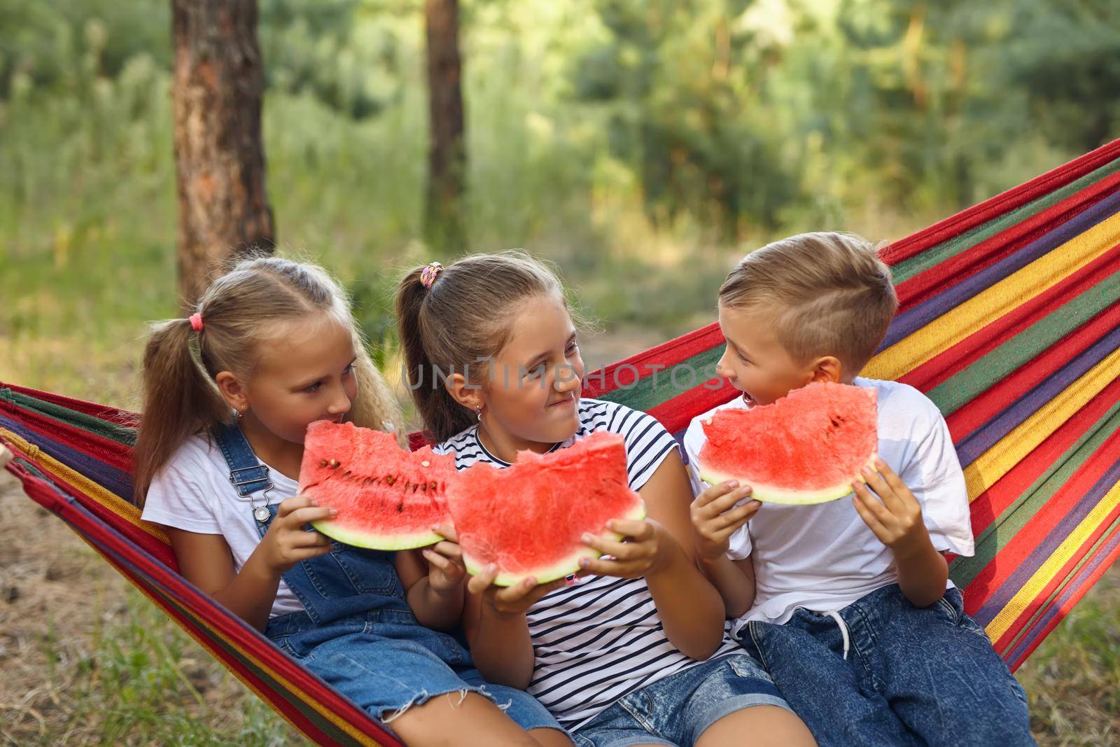 three cheerful children eat watermelon and joke, outdoor, sitting on a colorful hammock. Summer fun and leisure