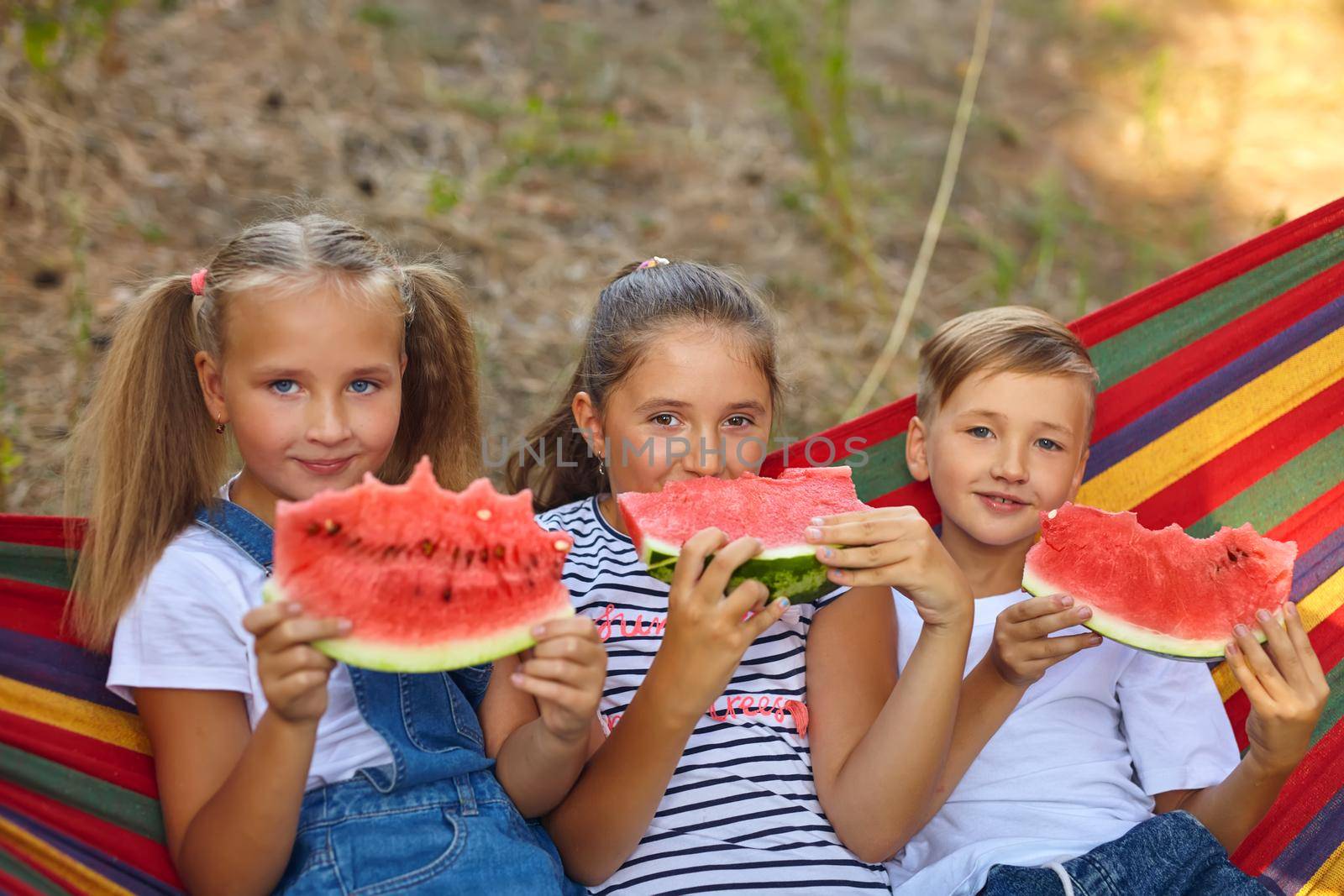 three cheerful children eat watermelon and joke, outdoor, sitting on a colorful hammock. Summer fun and leisure