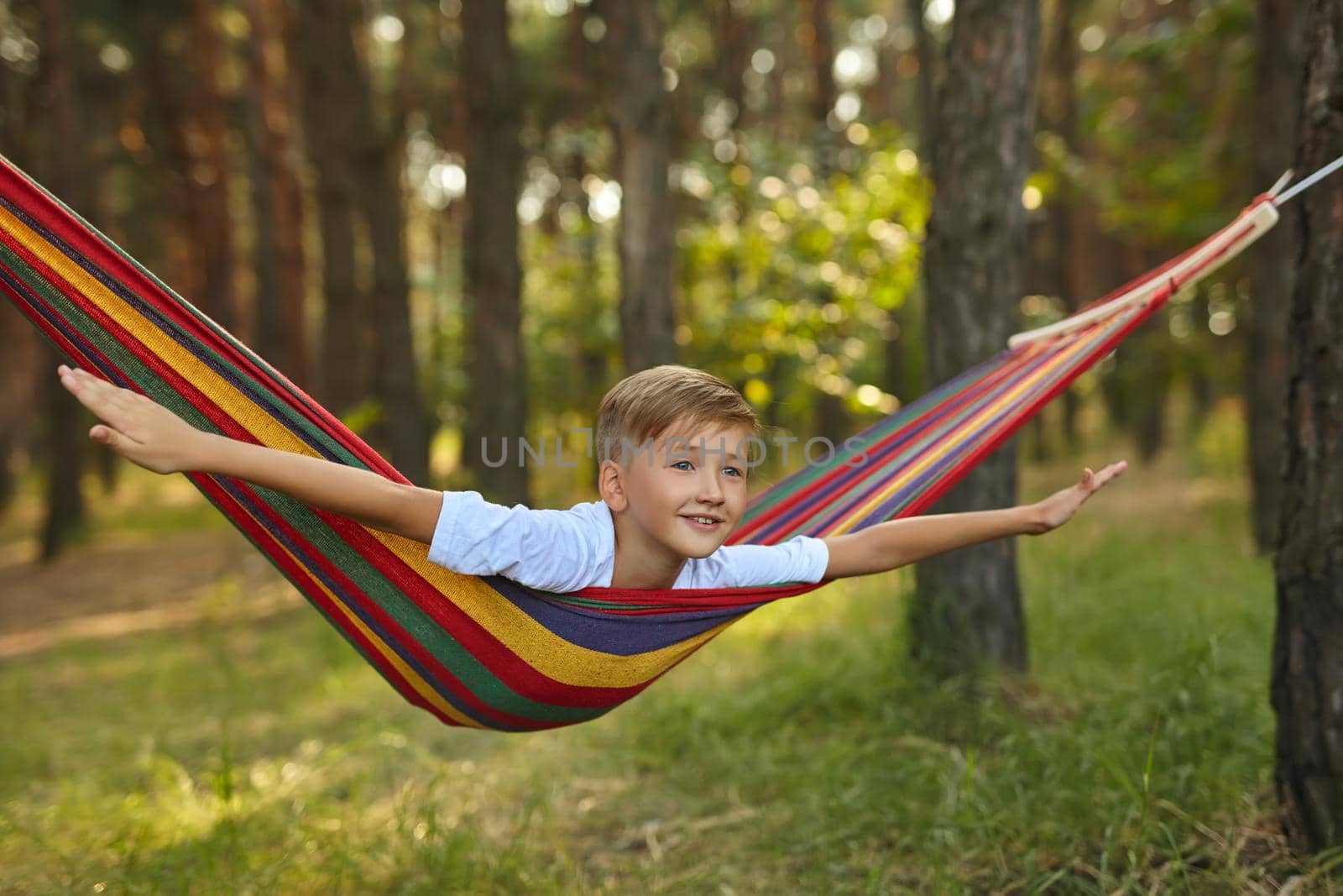 Cute little blond caucasian boy having fun with multicolored hammock in backyard or outdoor playground. Summer active leisure for kids. Child on hammock. Activities and fun for children outdoors